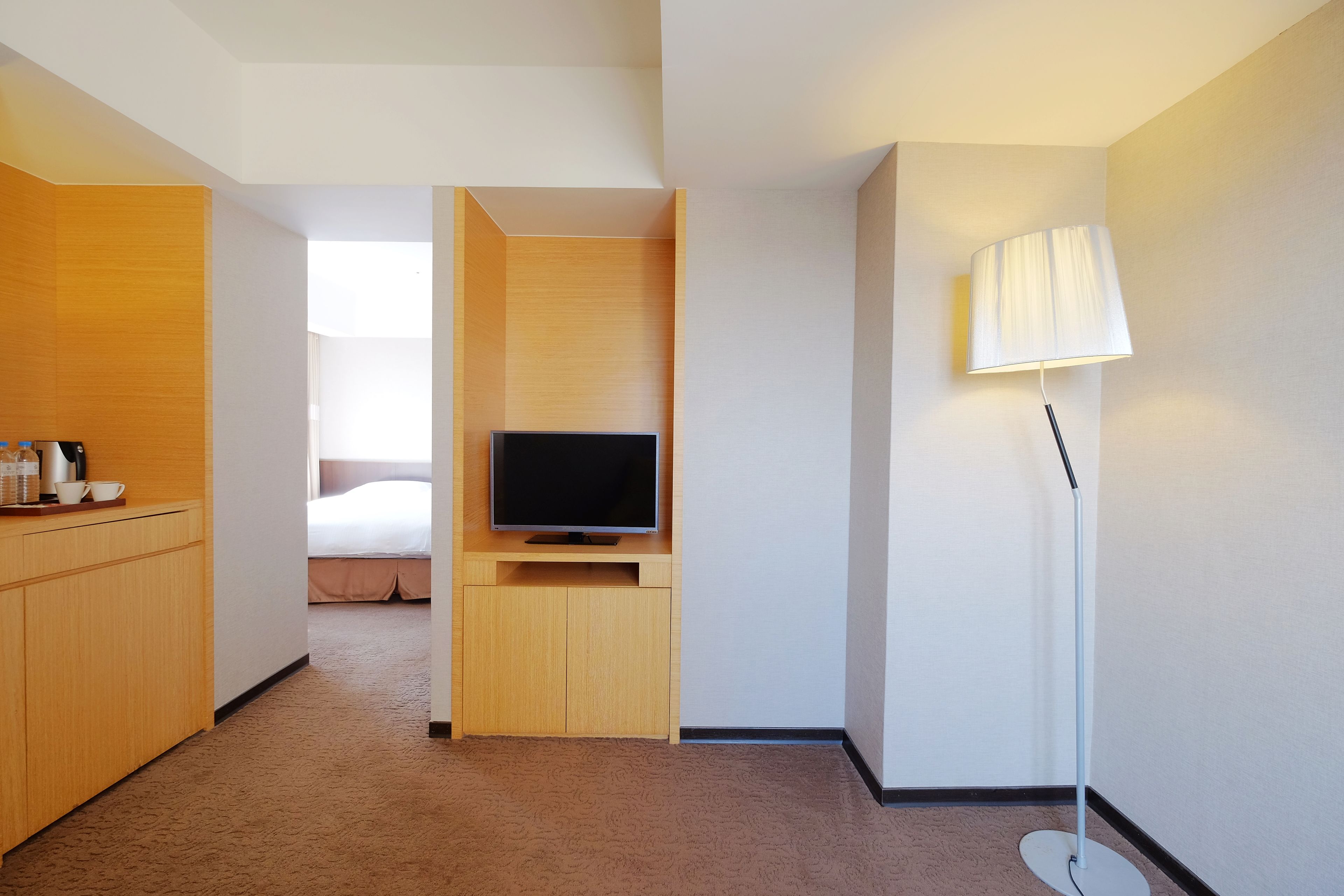 A hotel room with a wooden desk, a flat-screen TV, a standing lamp, and a bed in the background.