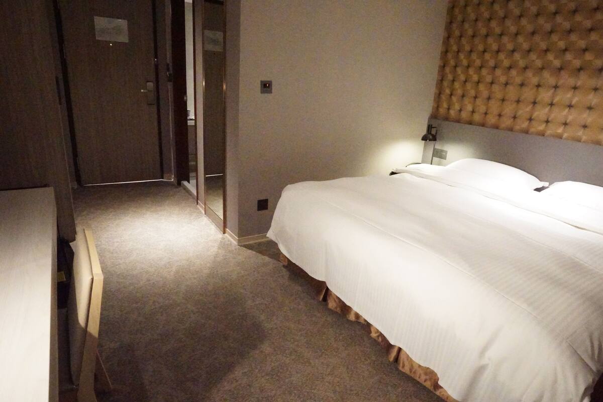 Een hotelkamer met een bed, een nachtkastje, een lamp en een deur naar een gang.