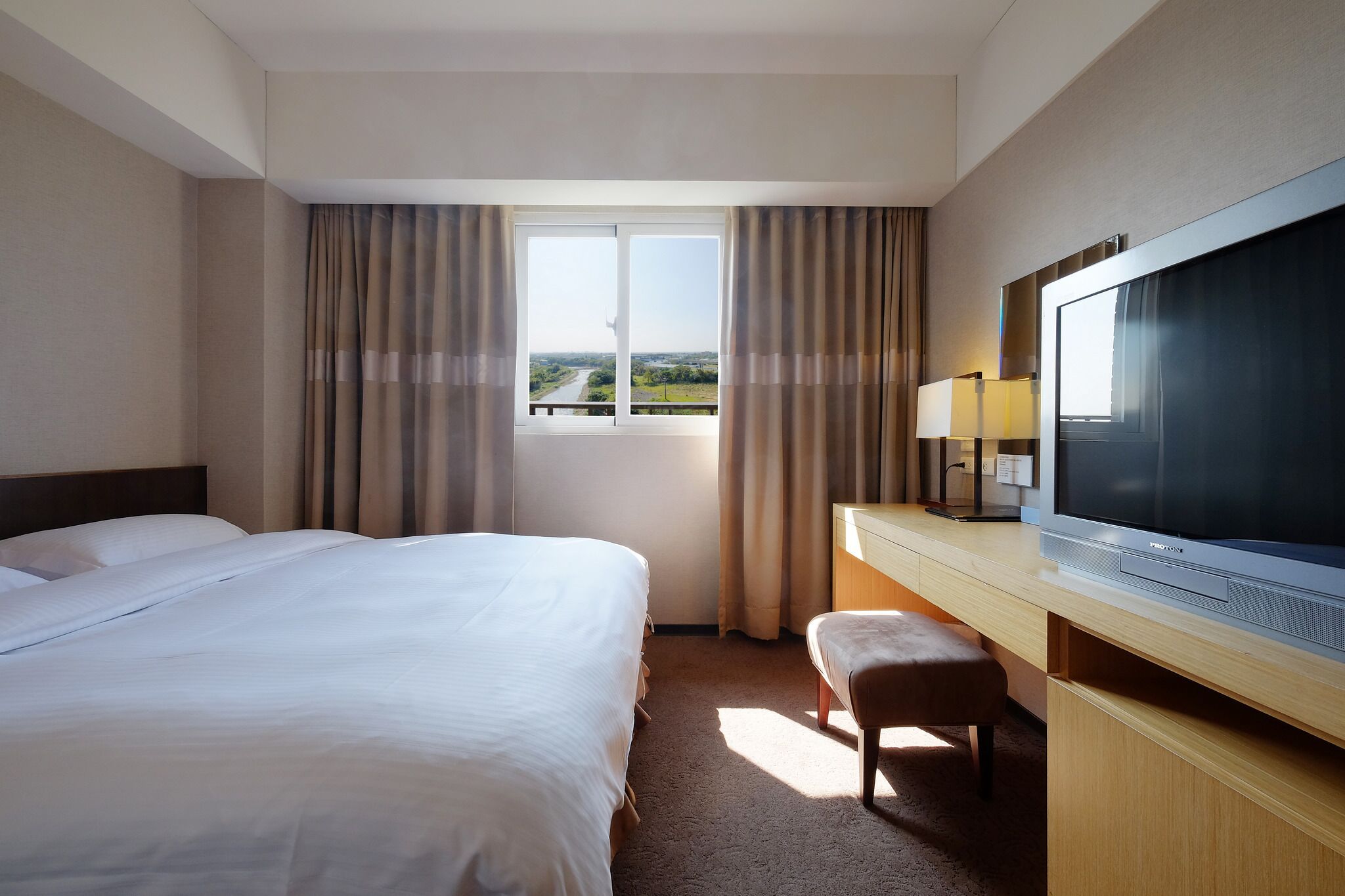 A hotel room with a large bed, a desk with a television, a window with curtains, and a view of a landscape.