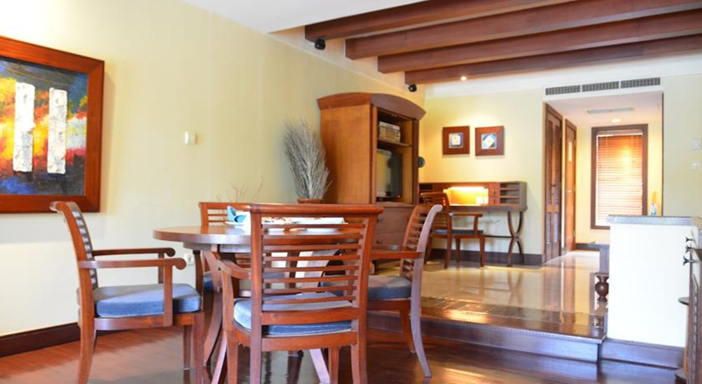 Un comedor con una mesa y sillas de madera, un cuadro en la pared y una cocina al fondo.