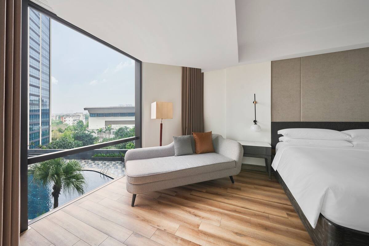 A modern hotel room with a large window, a sofa, a bed, and a nightstand.