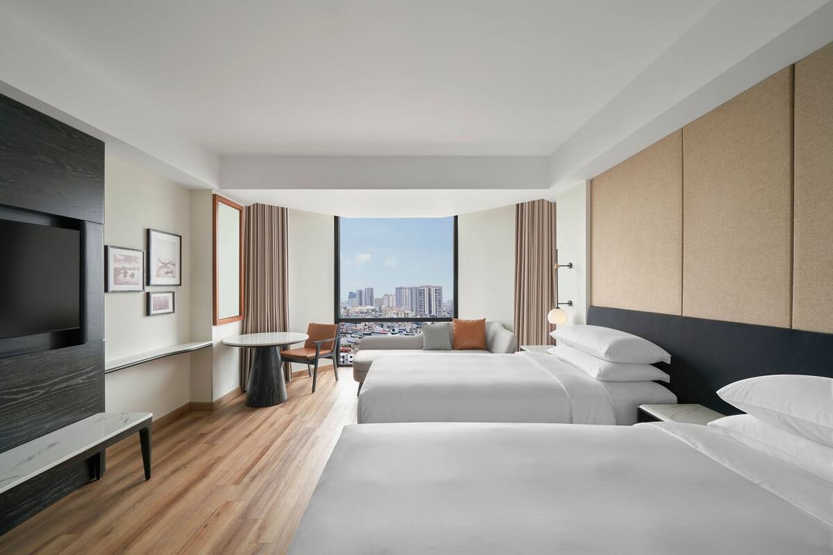 A modern hotel room with two beds, a TV, a dining table, and a city view.