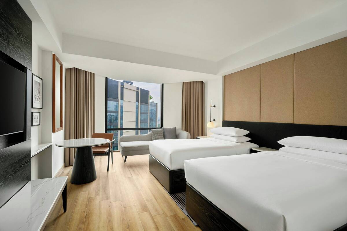 A modern hotel room with a large bed, a smaller bed, a desk, and a chair. There is a window with a view of buildings.