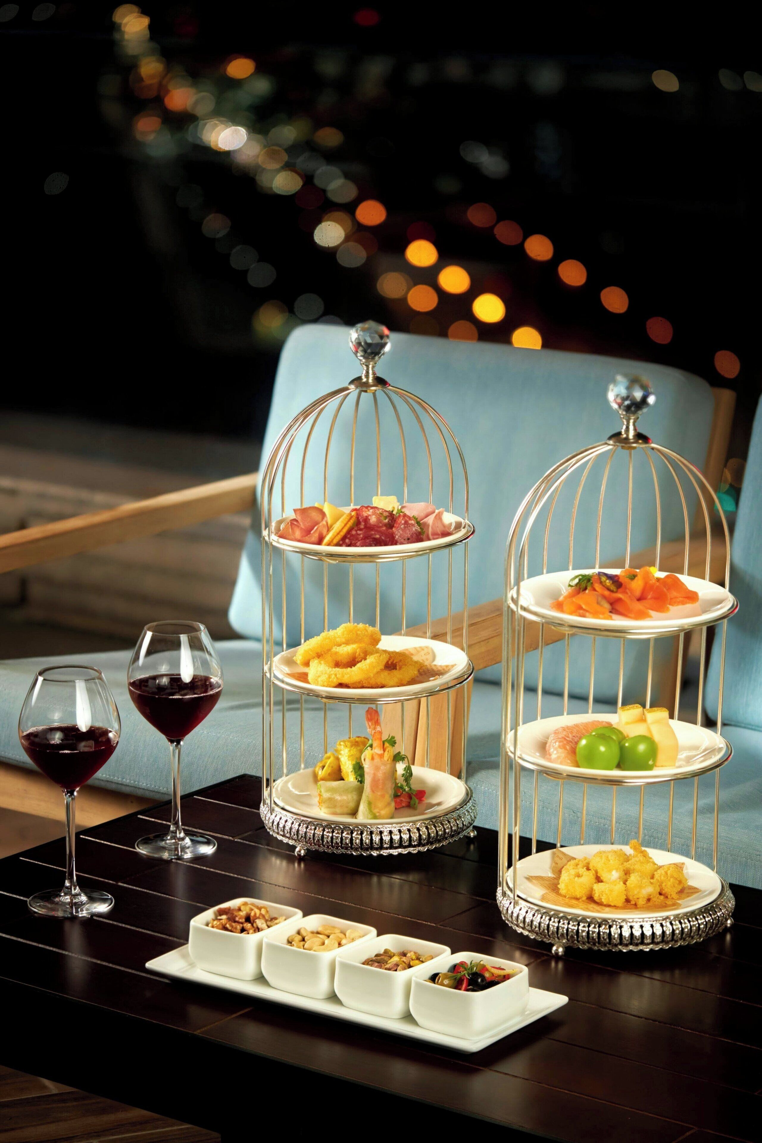 Un soporte de tres niveles para postres con diversos dulces y una bandeja de frutos secos, sobre una mesa con dos copas de vino.