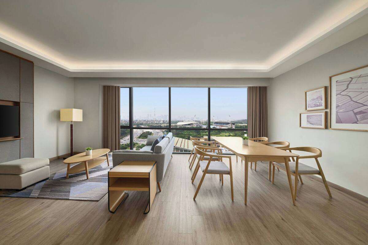 A modern hotel room with a large window offering a city view, a sofa, a dining table with chairs, and a flat-screen TV.