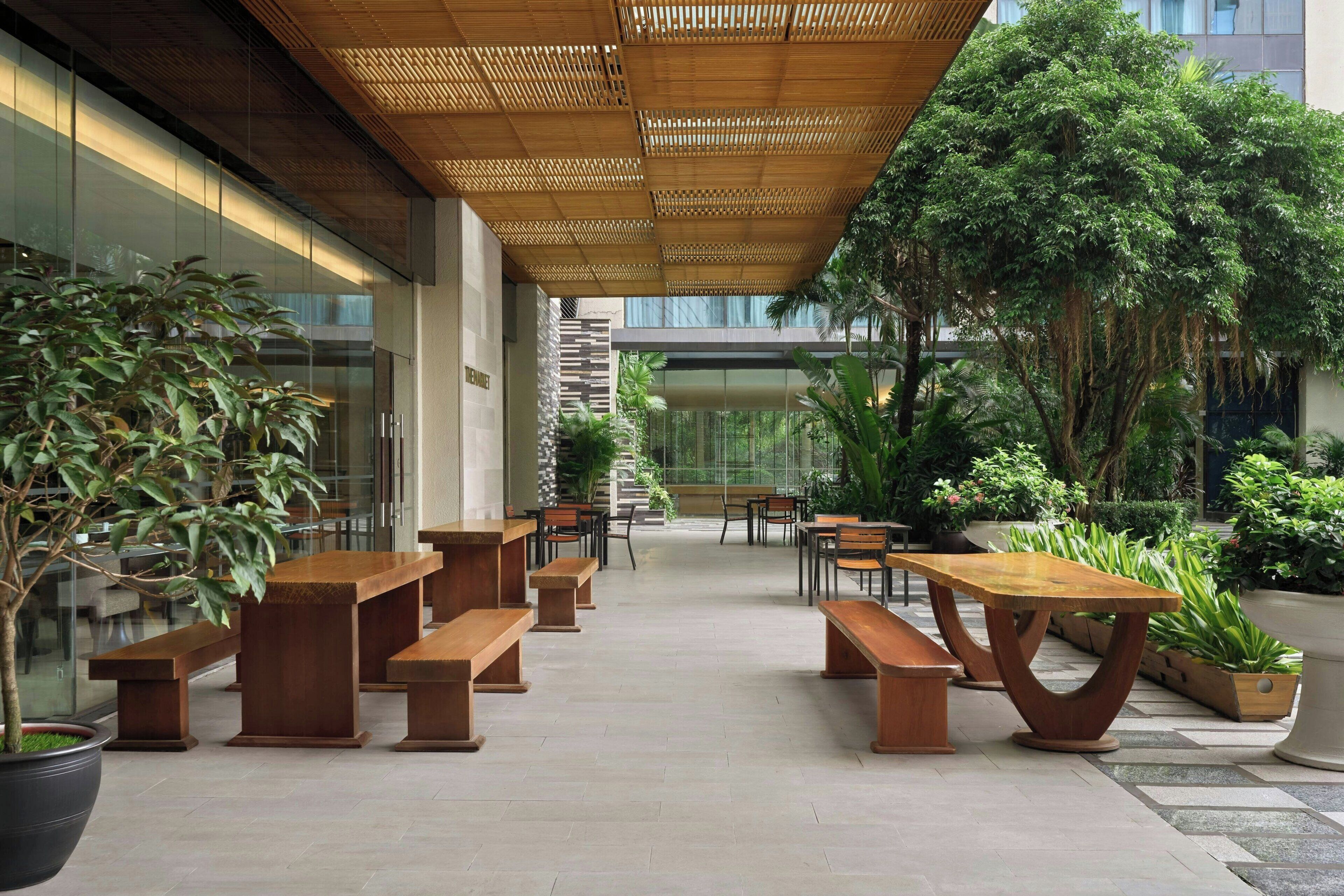 An outdoor patio with wooden benches and tables, surrounded by potted plants and trees.