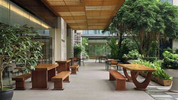 An outdoor patio with wooden benches and tables, surrounded by potted plants and trees.
