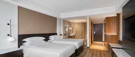 Habitación de hotel con dos camas, un escritorio, un televisor y un baño visible a través de una puerta abierta.