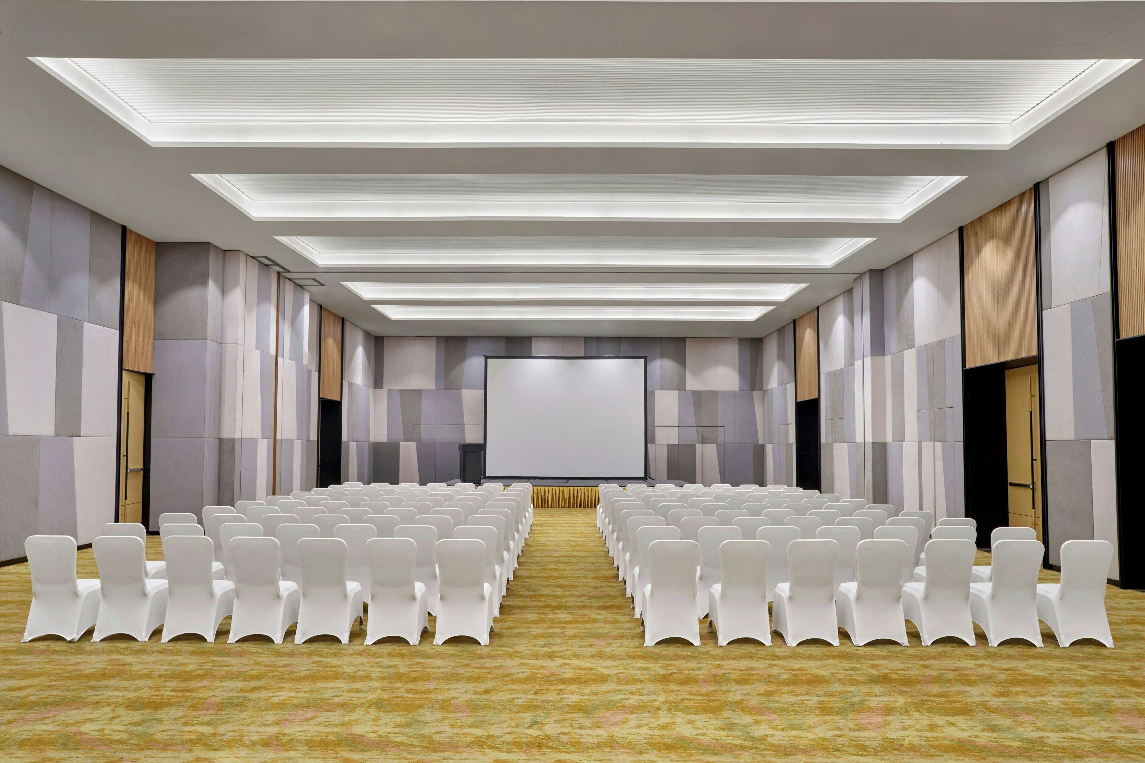 A conference room with rows of white chairs, a large screen at the front, and a patterned carpet.