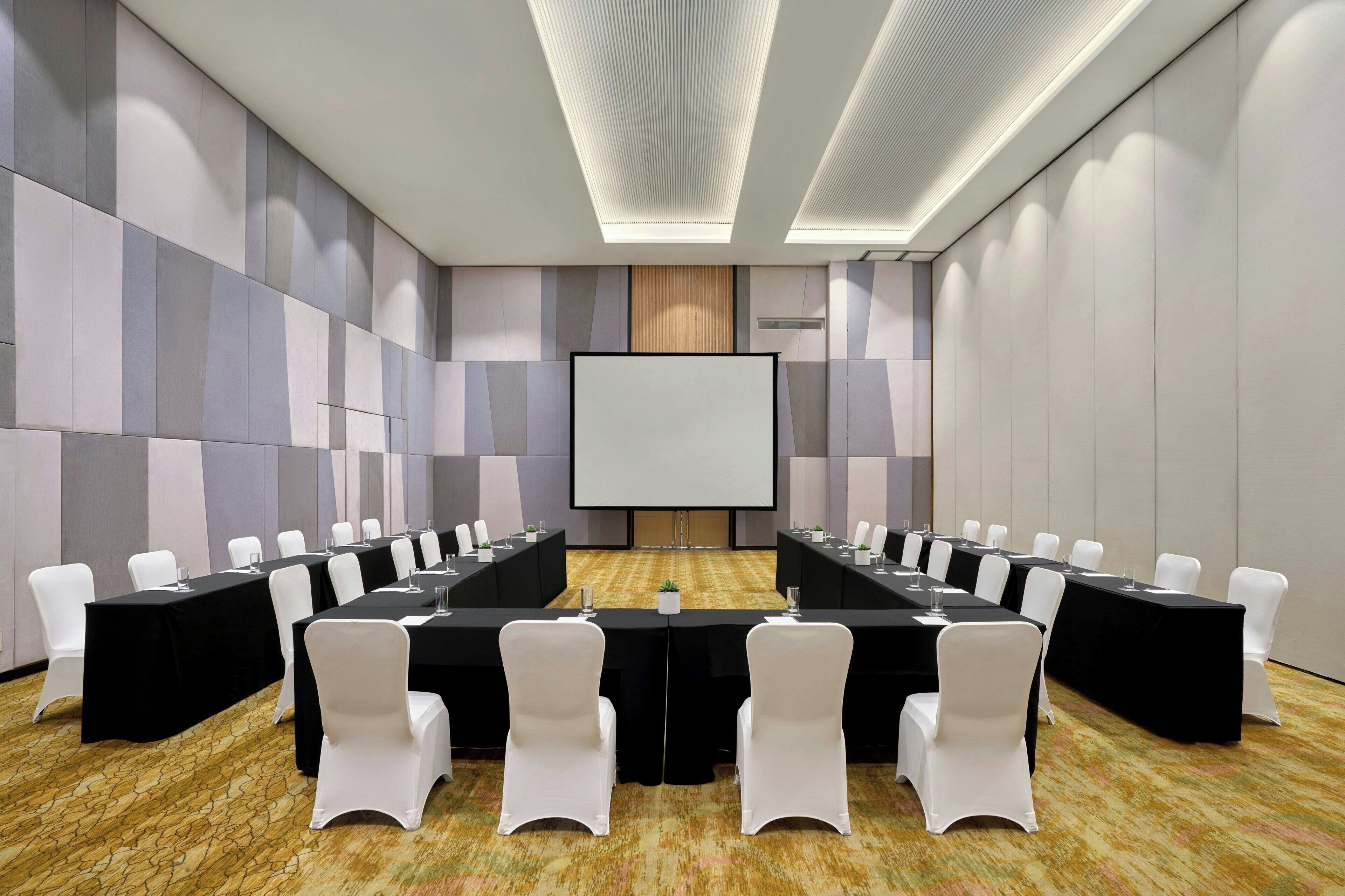 A conference room with a long table, white chairs, and a projector screen.
