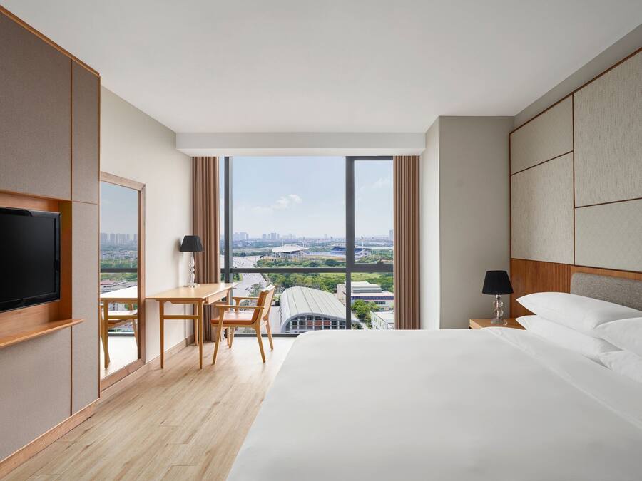 Habitación de hotel con una cama grande, un escritorio con una silla, un televisor y vistas de la ciudad a través de grandes ventanales.