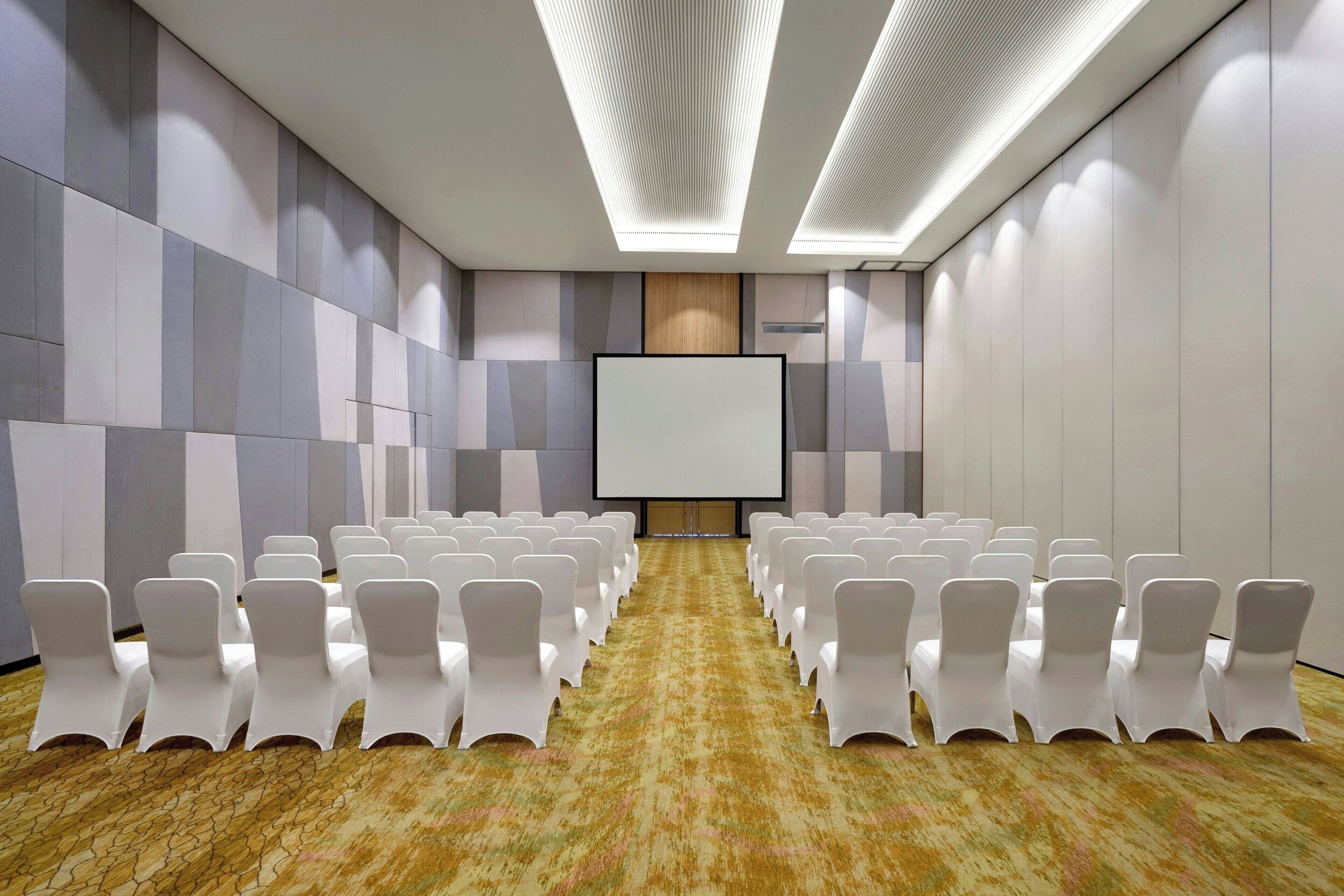 A conference room with rows of white chairs, a large screen, and a patterned carpet.