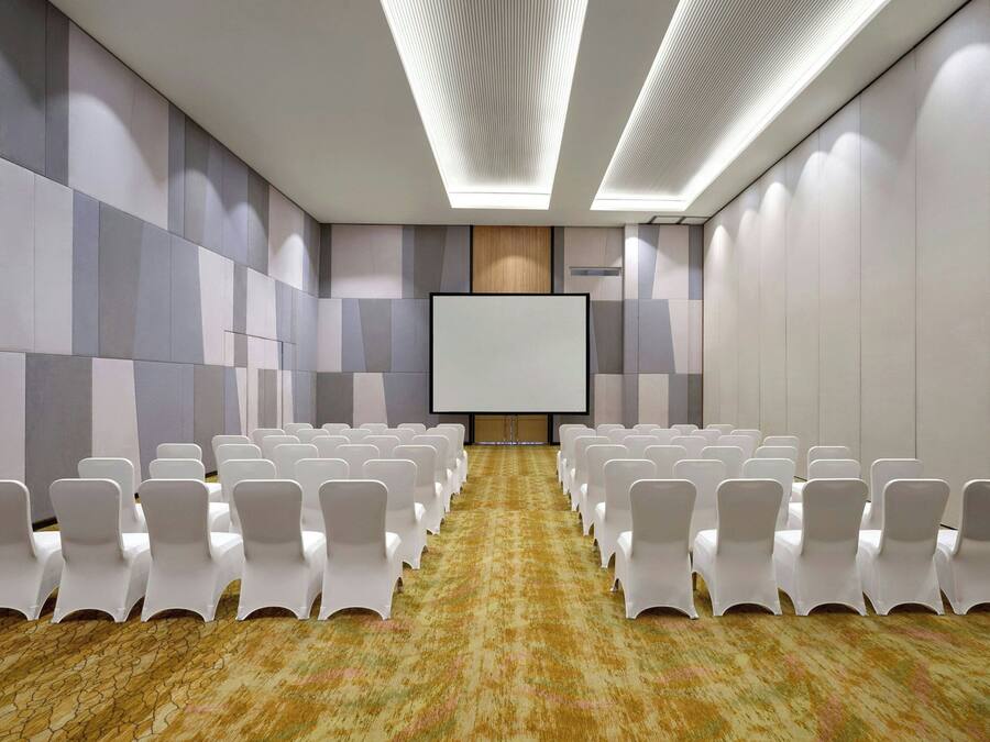 Un salón de conferencias con filas de sillas blancas, una pantalla grande y una alfombra estampada.
