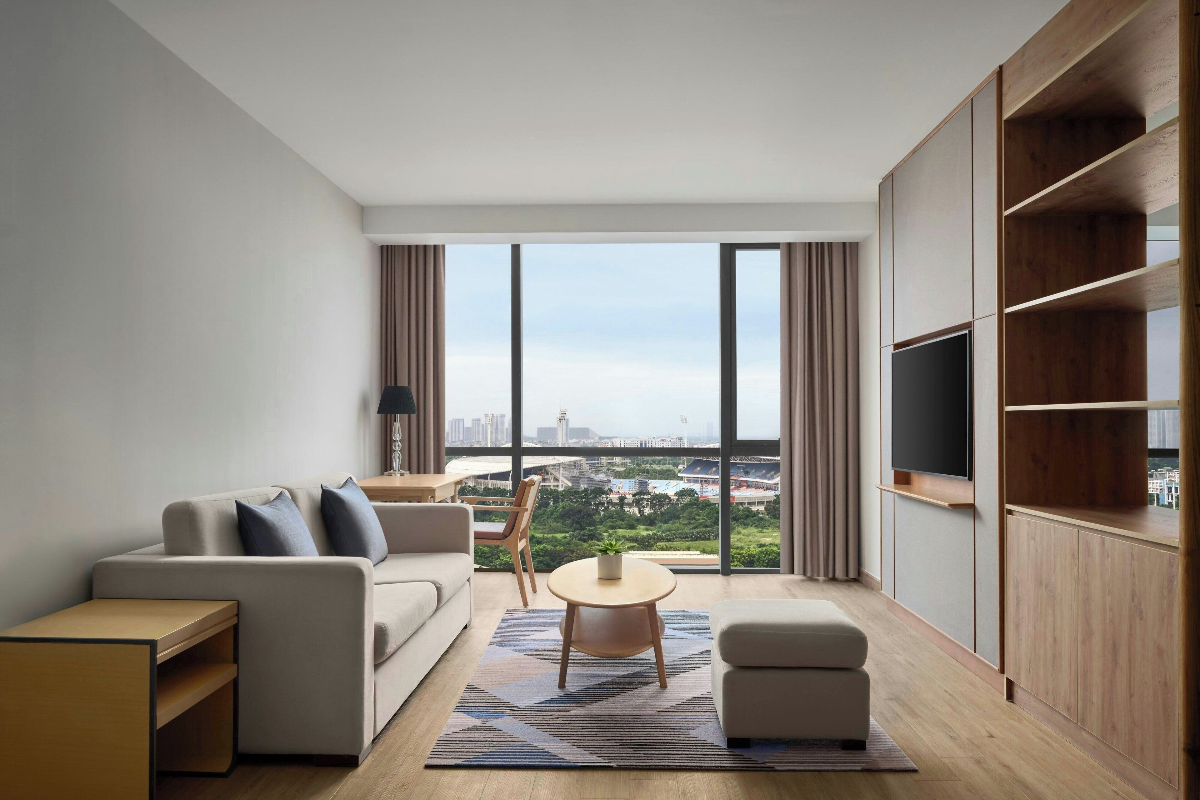 Una sala de estar moderna con un sofá, una mesita y un televisor. La habitación tiene ventanas amplias con vistas a un paisaje urbano.