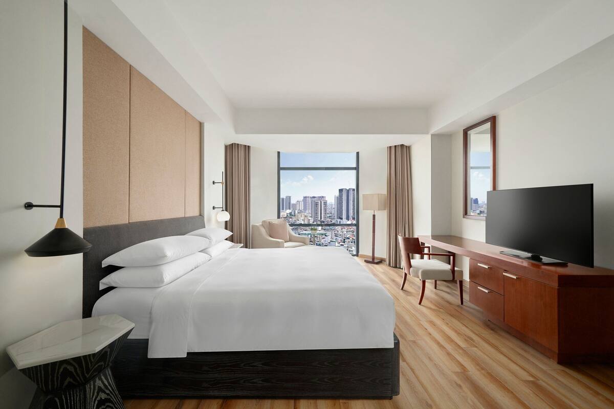 A hotel room with a large bed, a desk, a television, and a view of the cityscape.