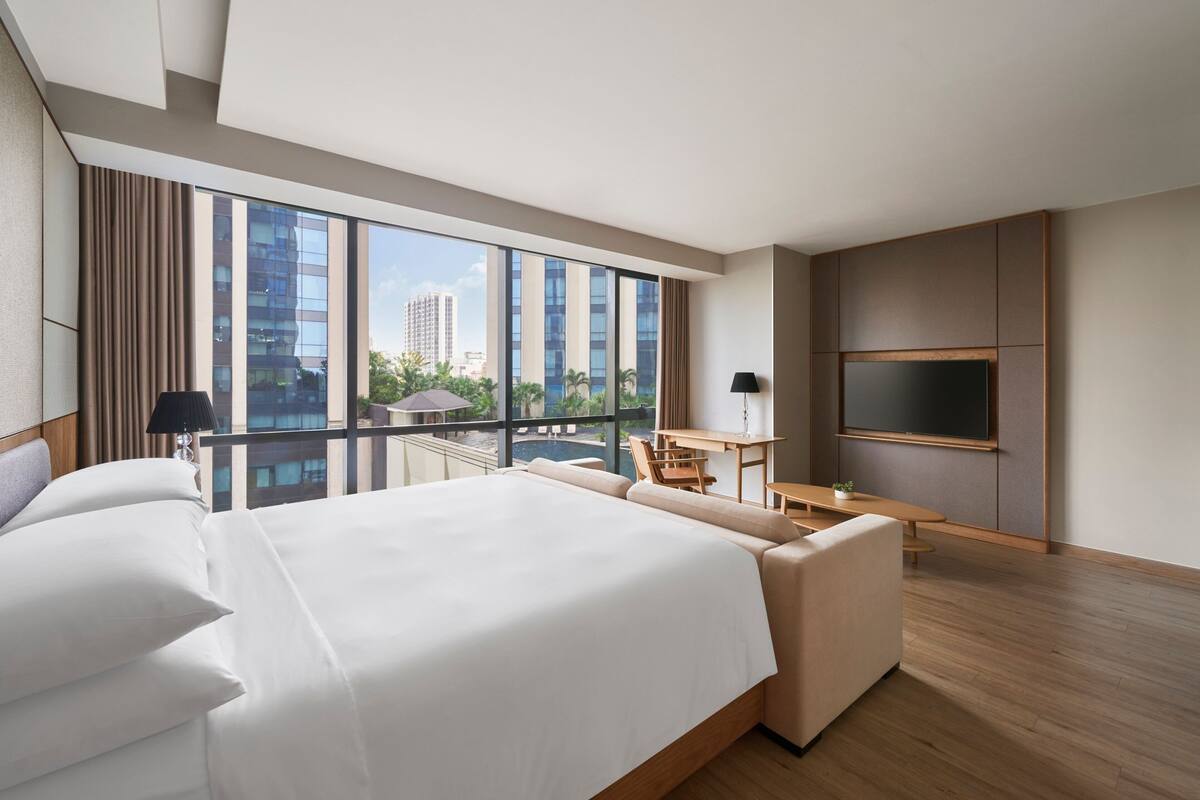 A modern hotel room with a large bed, a desk, a chair, a TV, and a balcony with a city view.