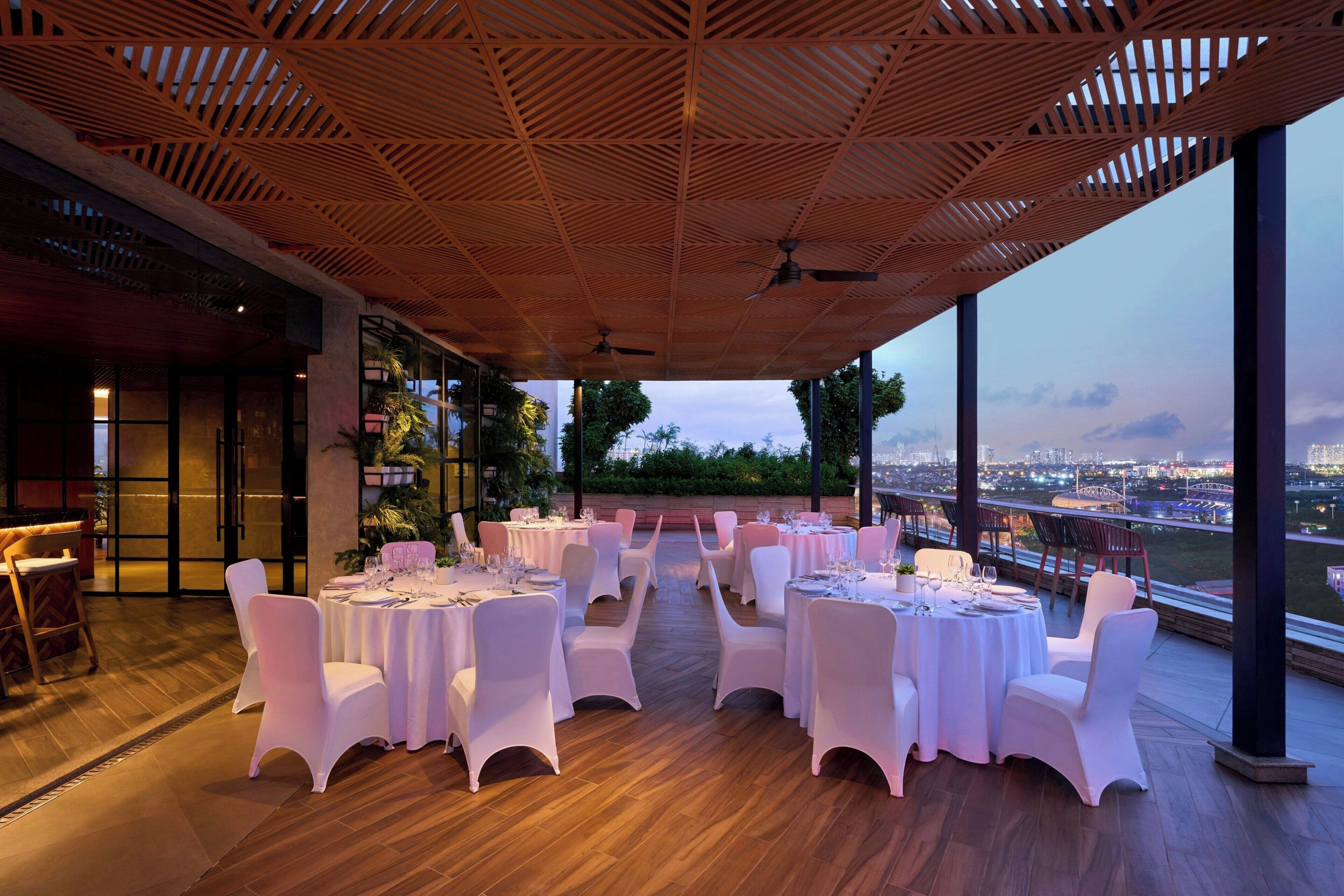 Un área de comedor al aire libre con mesas redondas y sillas blancas, con vistas a un paisaje urbano.