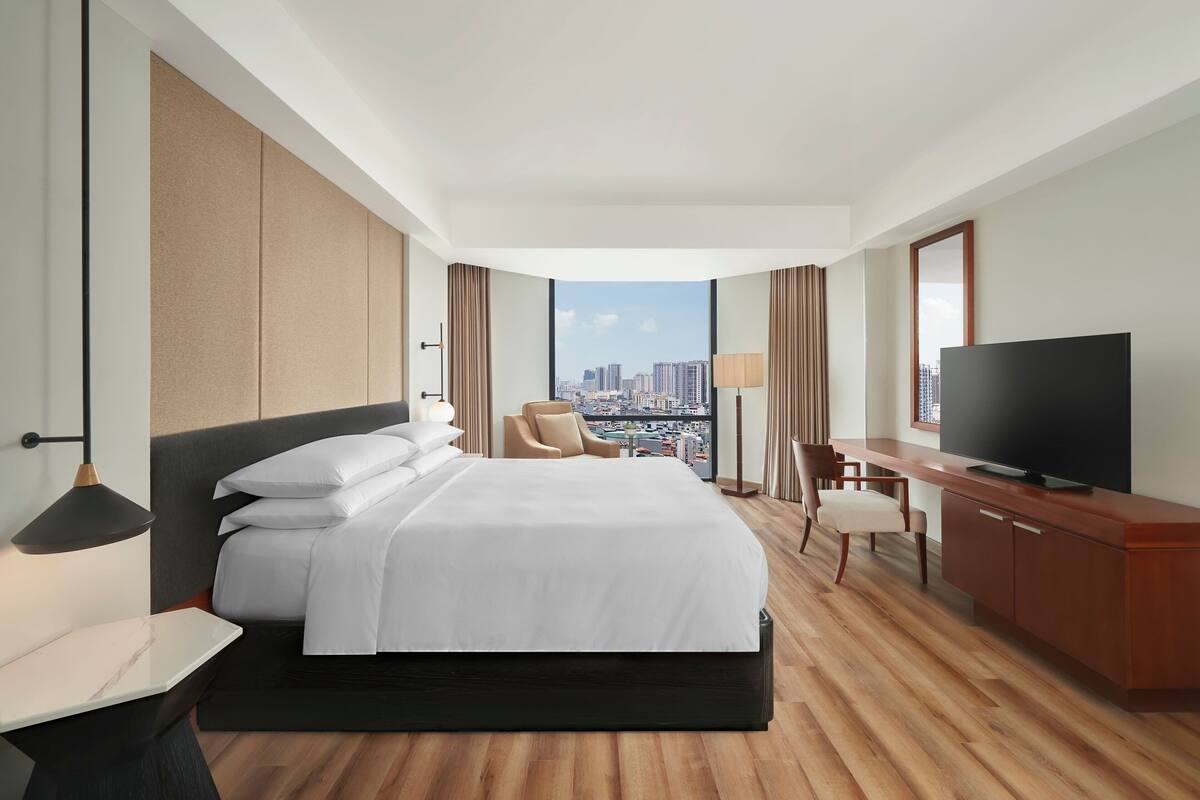 A hotel room with a large bed, a desk with a TV, a chair, and a view of the cityscape.