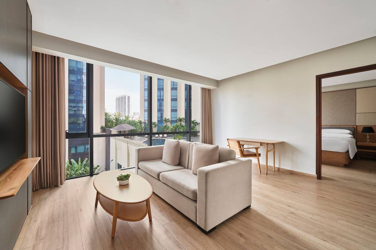 A modern hotel room with a large window, a sofa, a small table, a desk, and a bed.