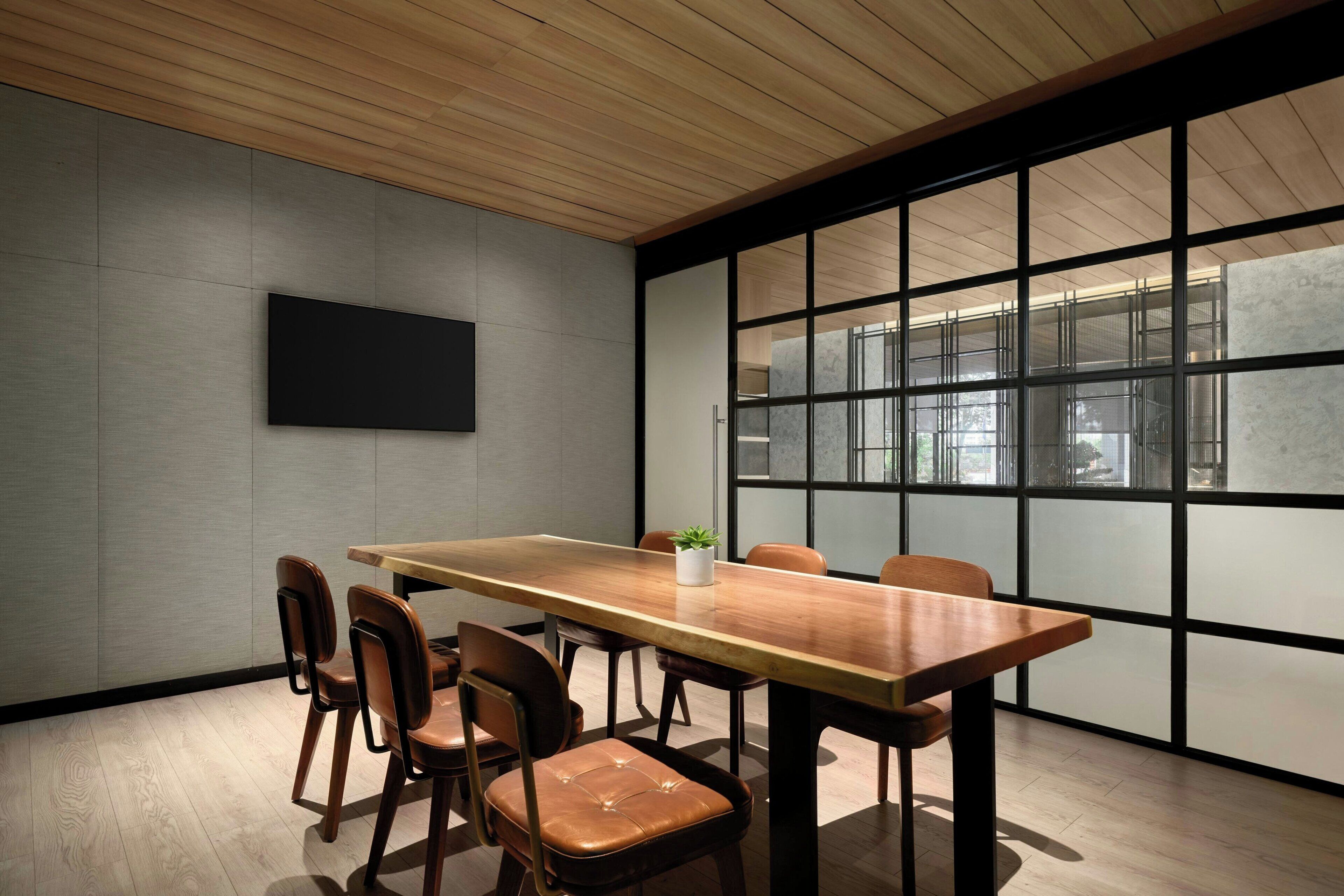 A conference room with a long wooden table, six chairs, a potted plant, and a large window with a view of buildings.