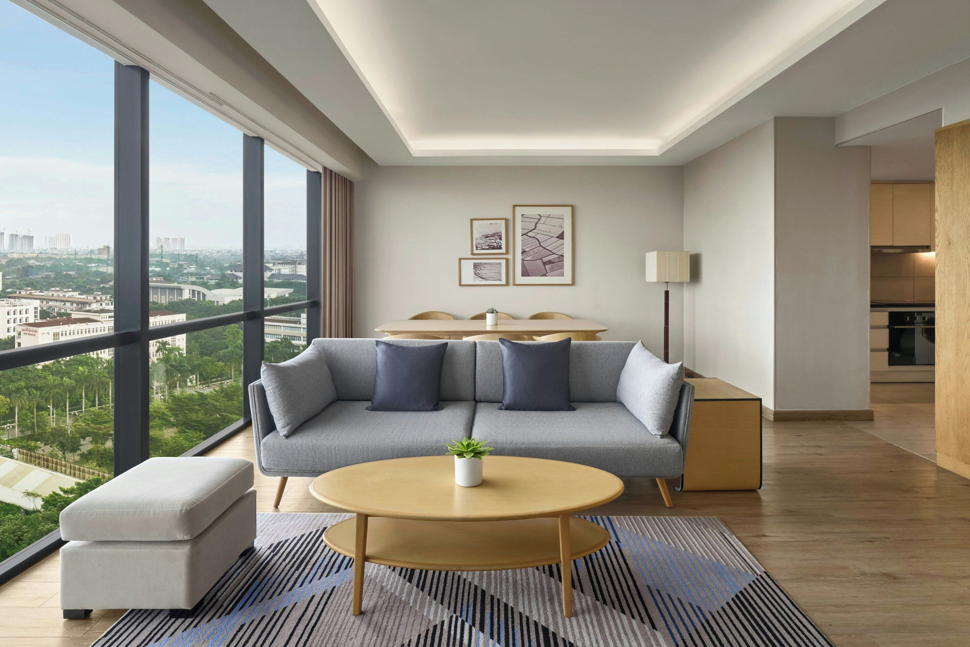 Una sala de estar moderna con un sofá, una mesa redonda de centro y vistas de la ciudad a través de grandes ventanales.