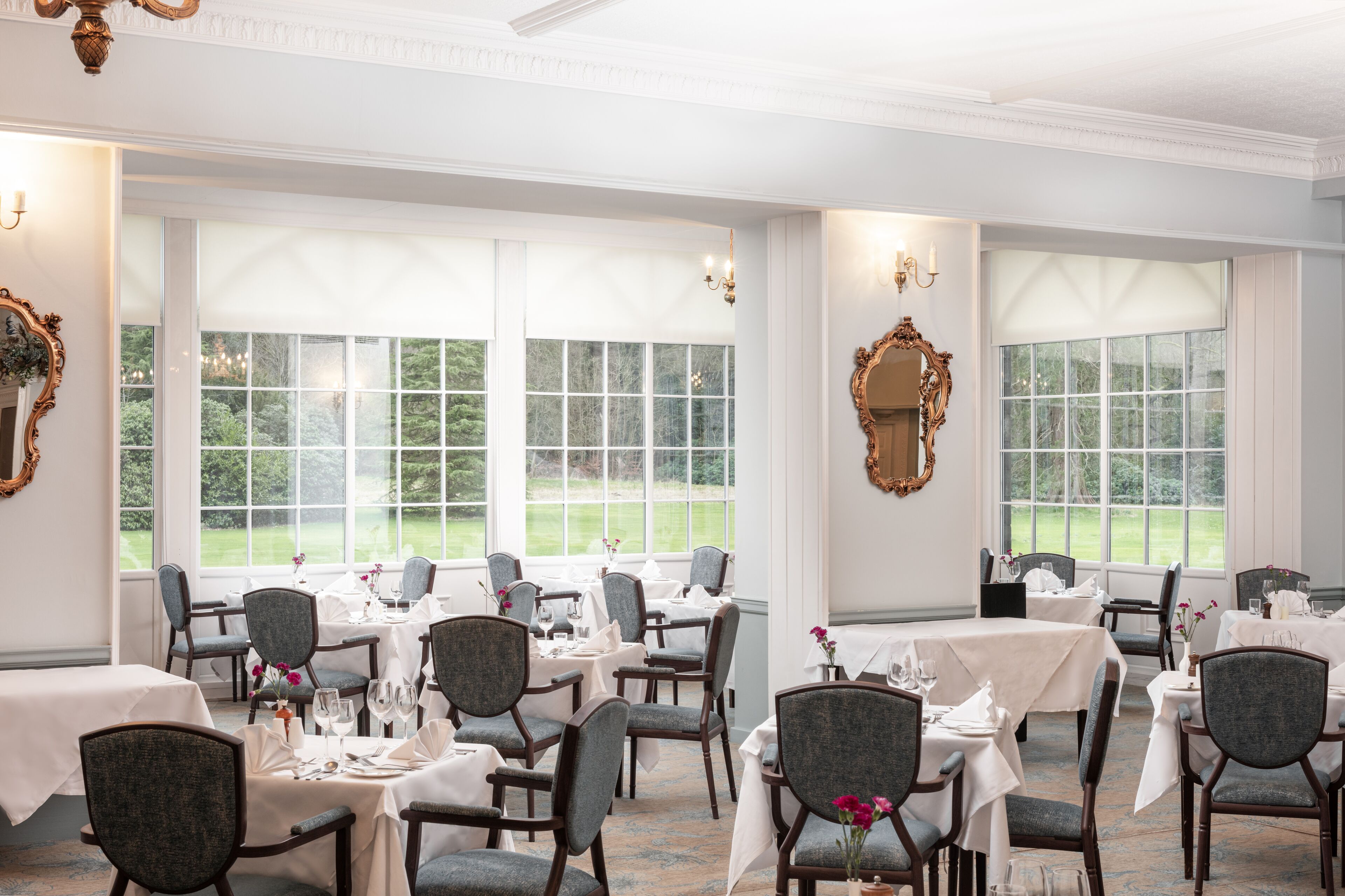 A dining area with tables set for service, large windows with a view of greenery, and ornate wall decorations.