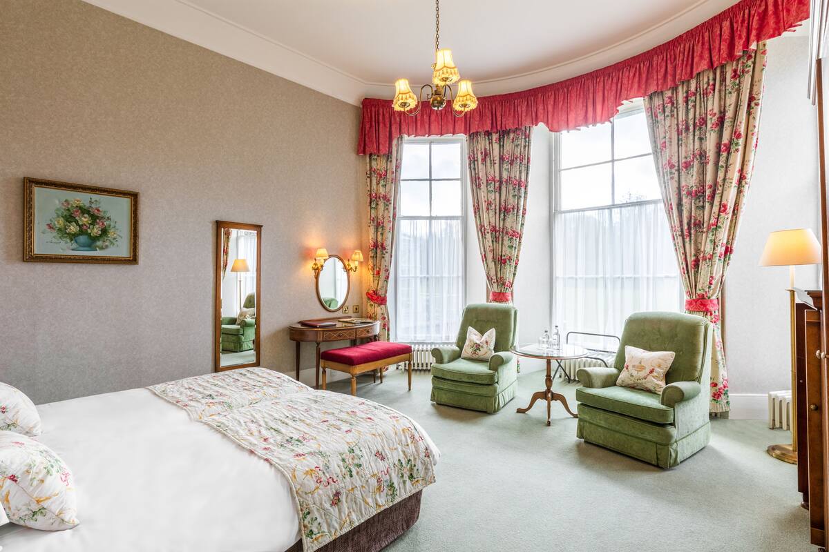 A bedroom with a large bed, two green armchairs, a small table, a mirror, and floral curtains.