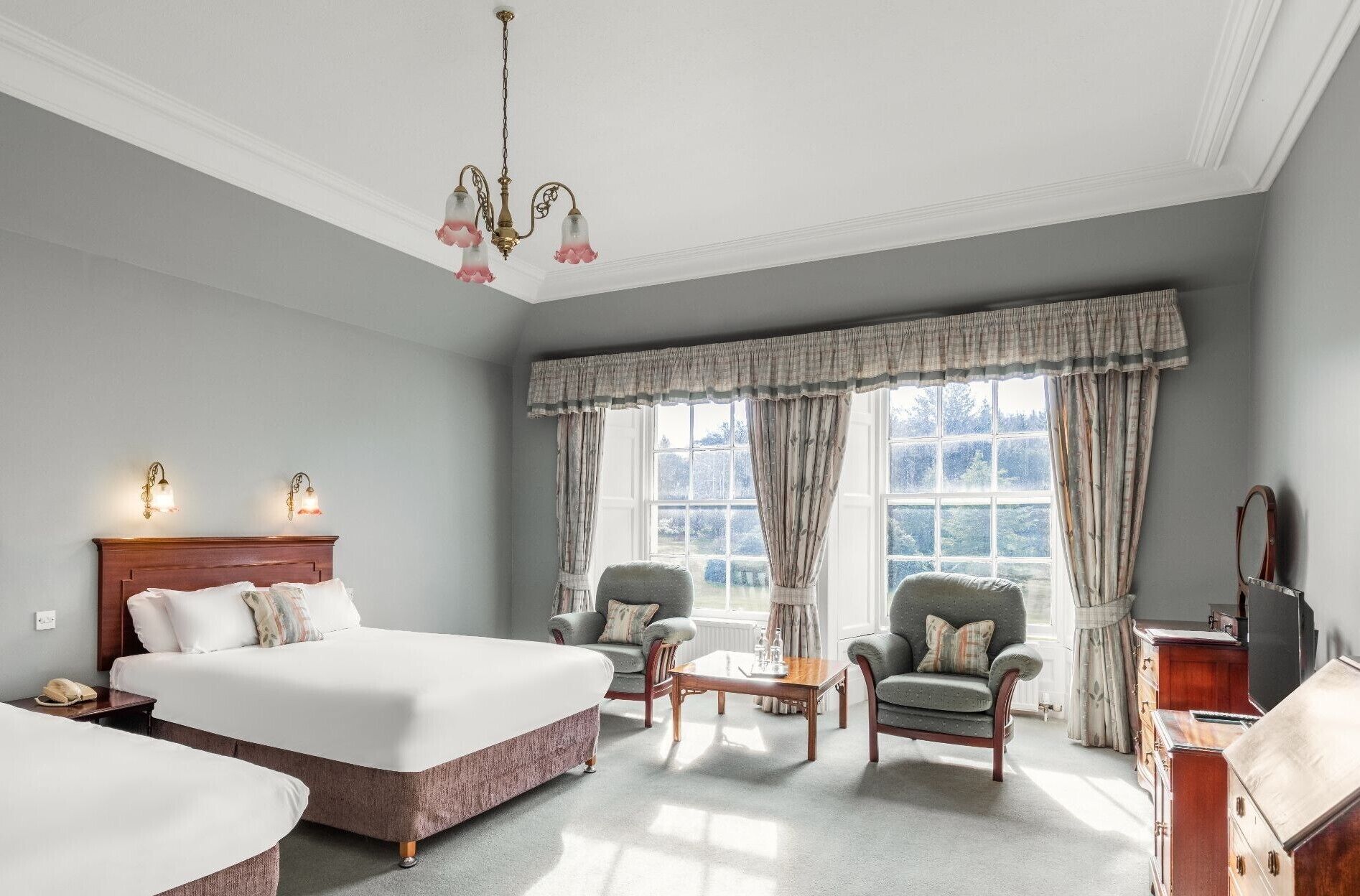 A hotel room with a large bed, two armchairs, a small table, a wooden dresser, and a large window with curtains.