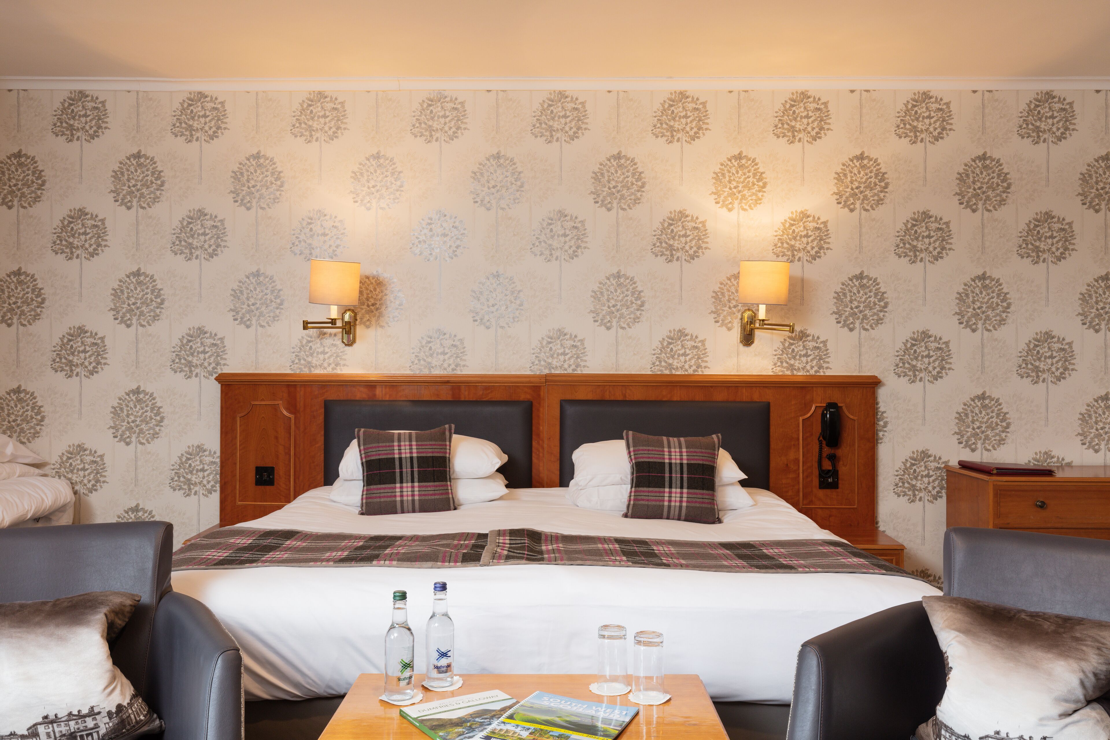 A hotel room with a large bed, two bedside lamps, a wooden headboard, and a table with a magazine and glasses.