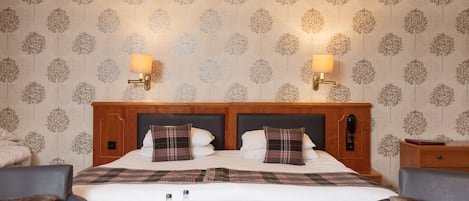 A hotel room with a large bed, two bedside lamps, a wooden headboard, and a table with a magazine and glasses.