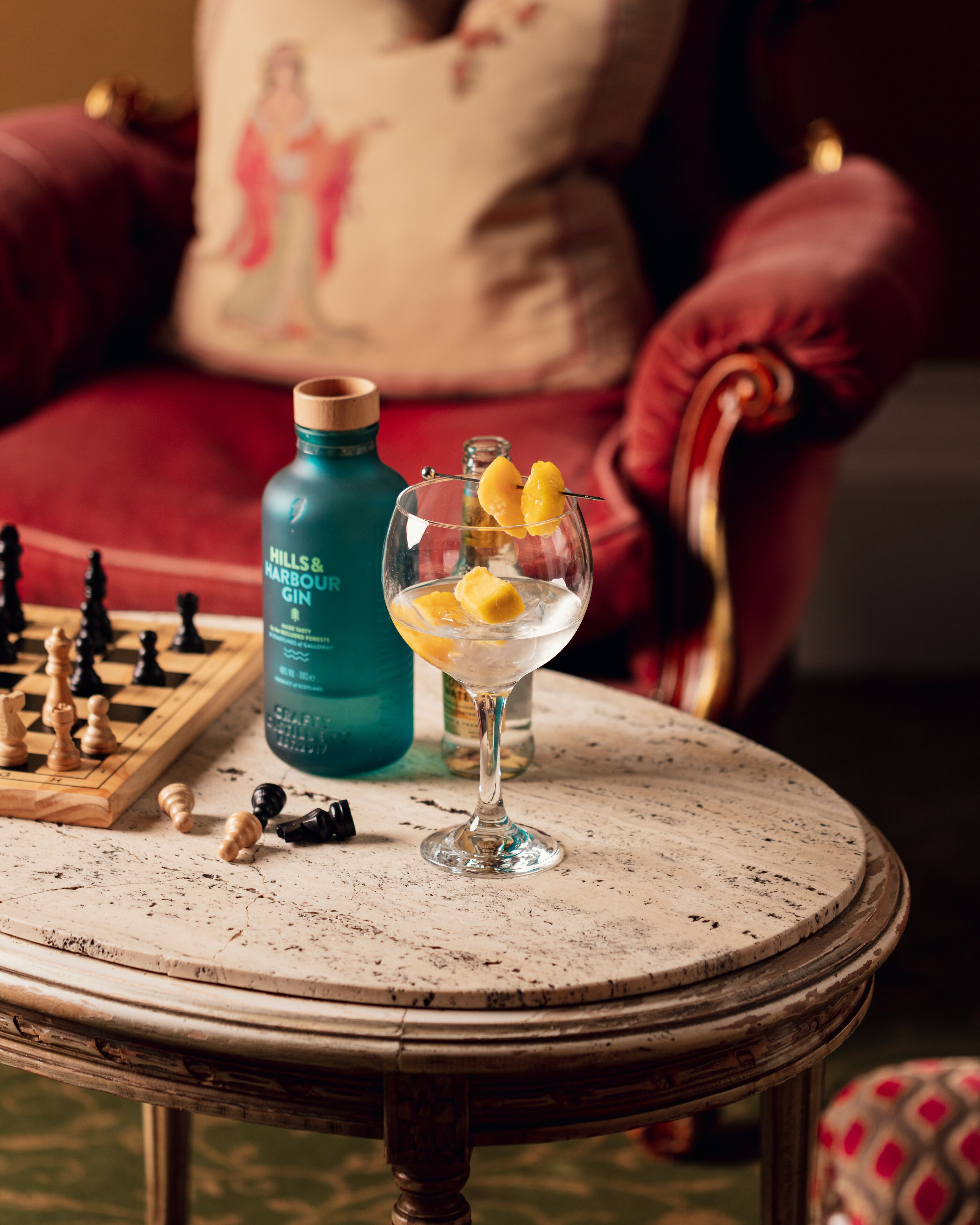 A round table with a glass of gin and lemon slices, a chessboard, and a bottle of gin.