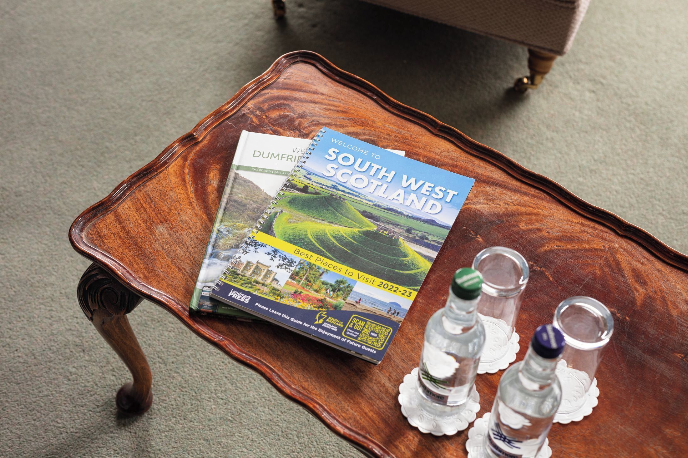 A wooden tray with a travel guide, two water bottles, and a glass on a carpeted floor.