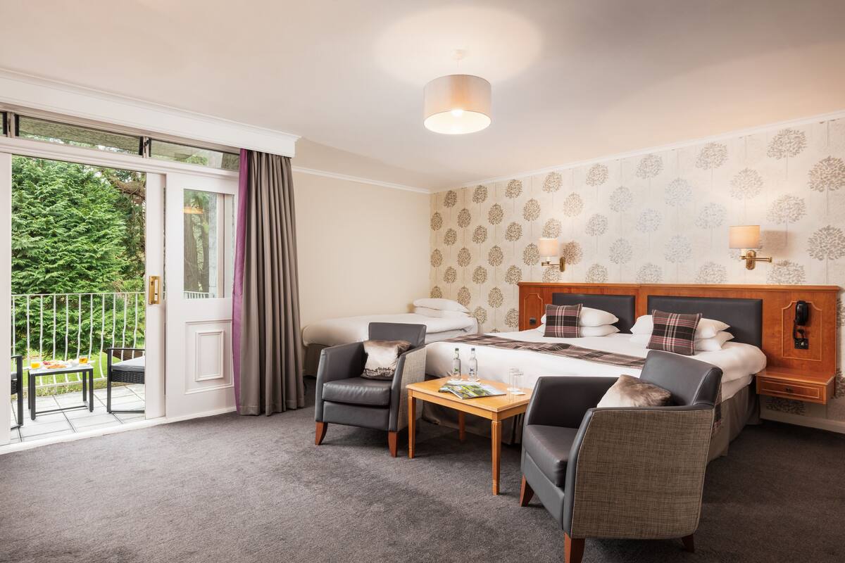 A hotel room with a bed, two armchairs, a small table, a nightstand, and a balcony with a view of greenery.