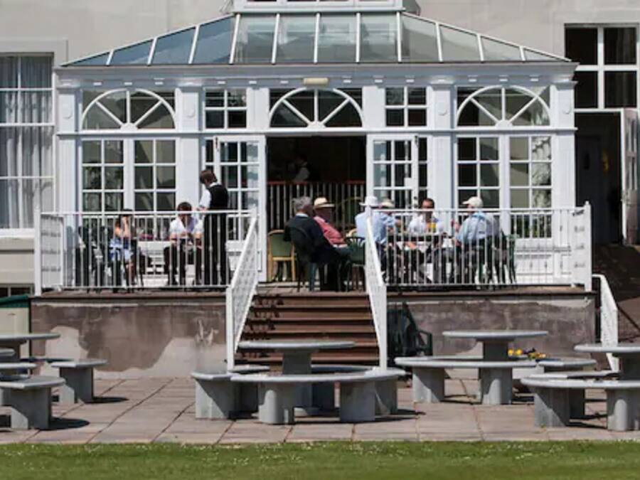 A covered outdoor seating area with tables and chairs, a staircase leading to an entrance, and a building with large windows and a glass roof.