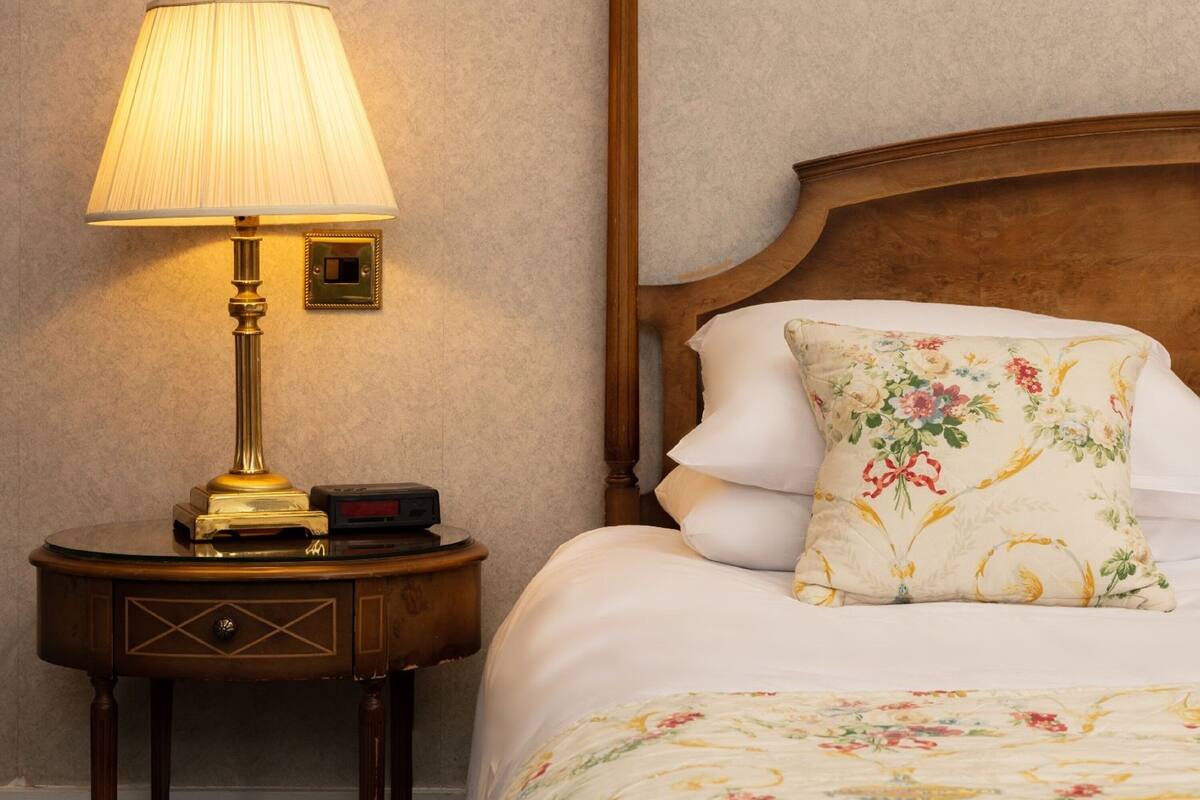 Uma cama bem arrumada com roupa de cama floral, cabeceira de madeira e mesa de cabeceira com abajur e relógio.