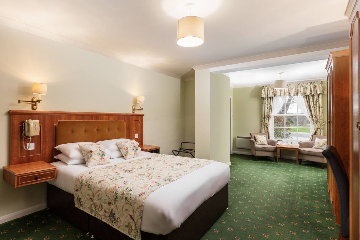 A hotel room with a large bed, bedside lamps, a telephone, a small table, two chairs, and a window with curtains.