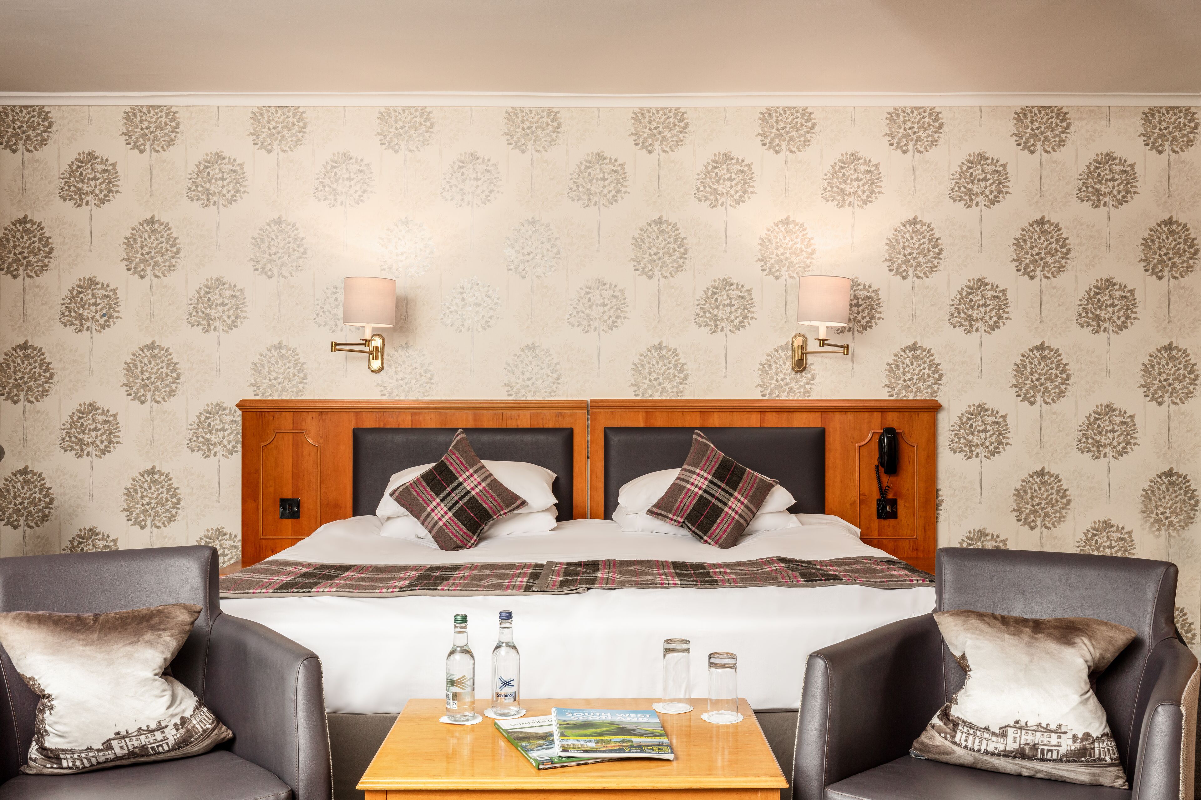 A hotel room with a double bed, two bedside tables, two chairs, a wooden headboard, floral wallpaper, and two wall-mounted lamps.