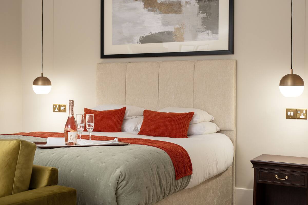 A neatly made bed with a red and white color scheme, a bedside table with a bottle and glasses, and a framed artwork above the bed.