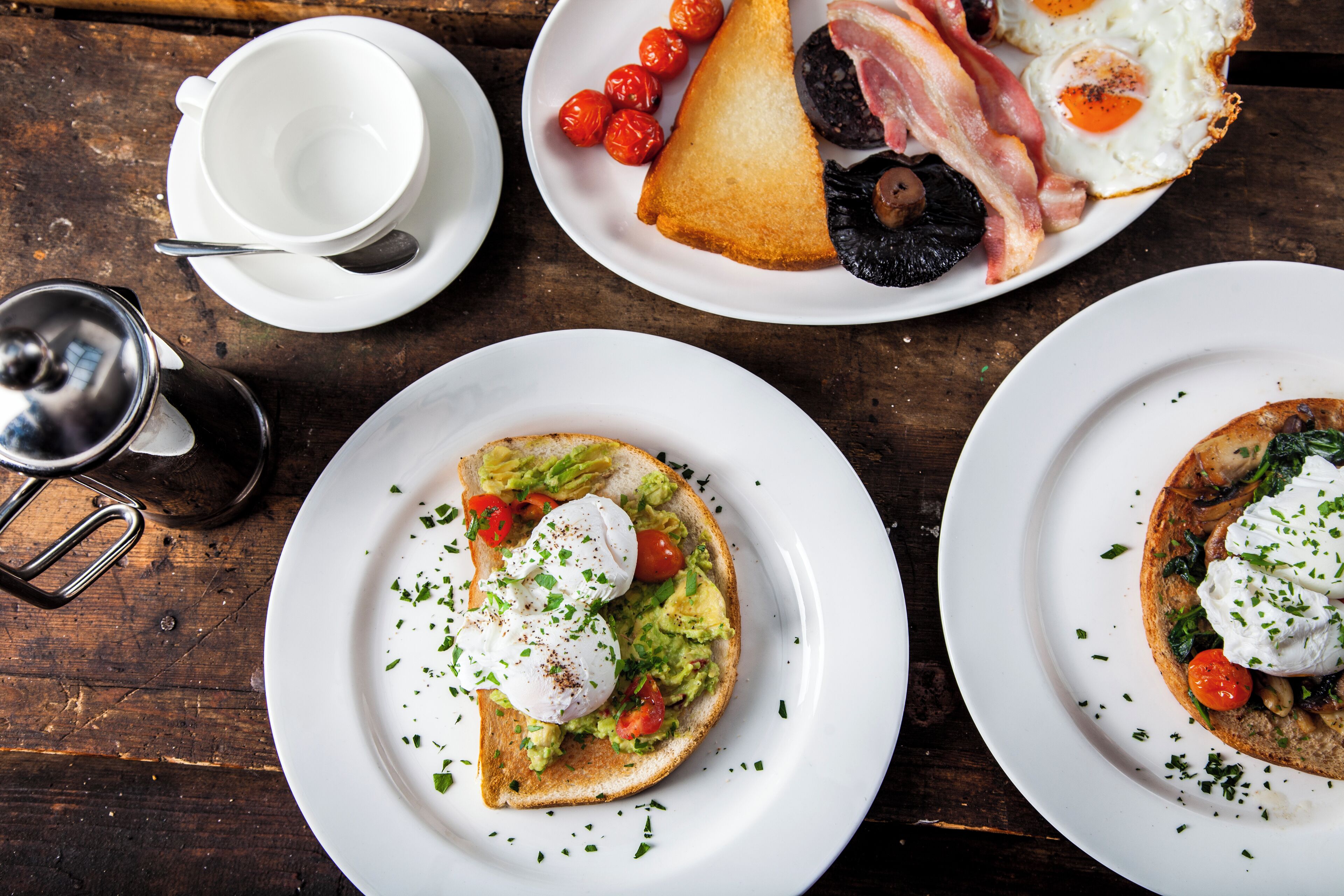 Une table en bois sur laquelle sont disposées des assiettes contenant des aliments pour le petit-déjeuner, notamment des toasts à l’avocat et des œufs pochés, ainsi qu’une assiette avec du bacon, des œufs et des tomates.