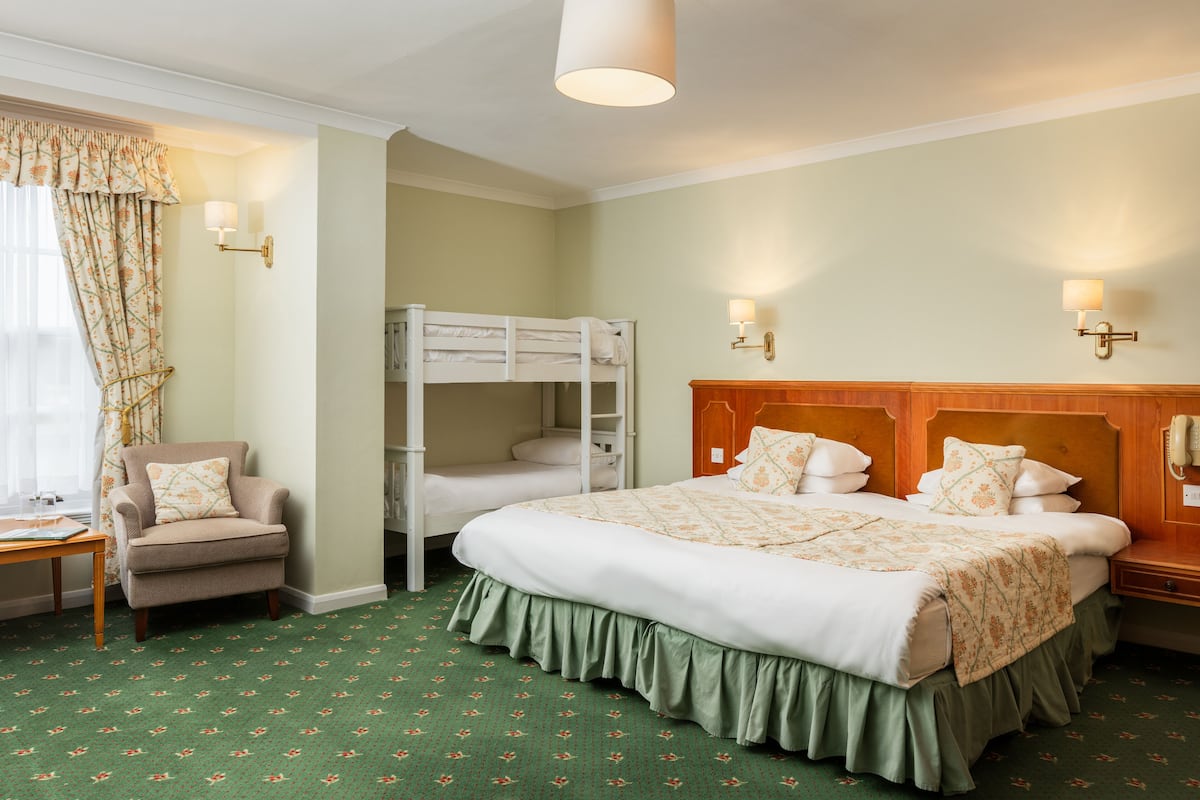 A hotel room with a bunk bed, a single bed, a chair, a table, and a lamp.