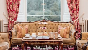A three-tiered tea service on a wooden table in front of a sofa with floral curtains. - Cally Palace Hotel & Golf Course (Castle Douglas)