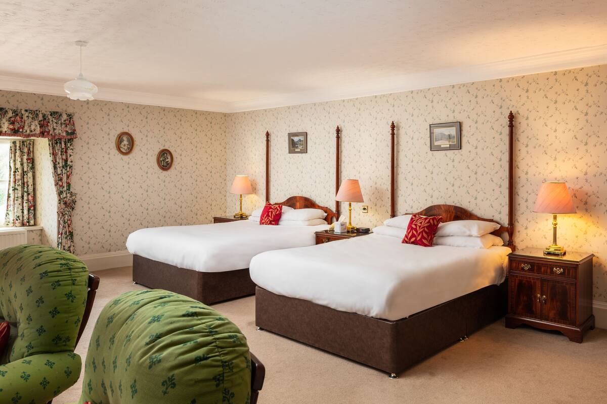 A hotel room with two beds, floral wallpaper, a wooden headboard, a nightstand with a lamp, and a green armchair.