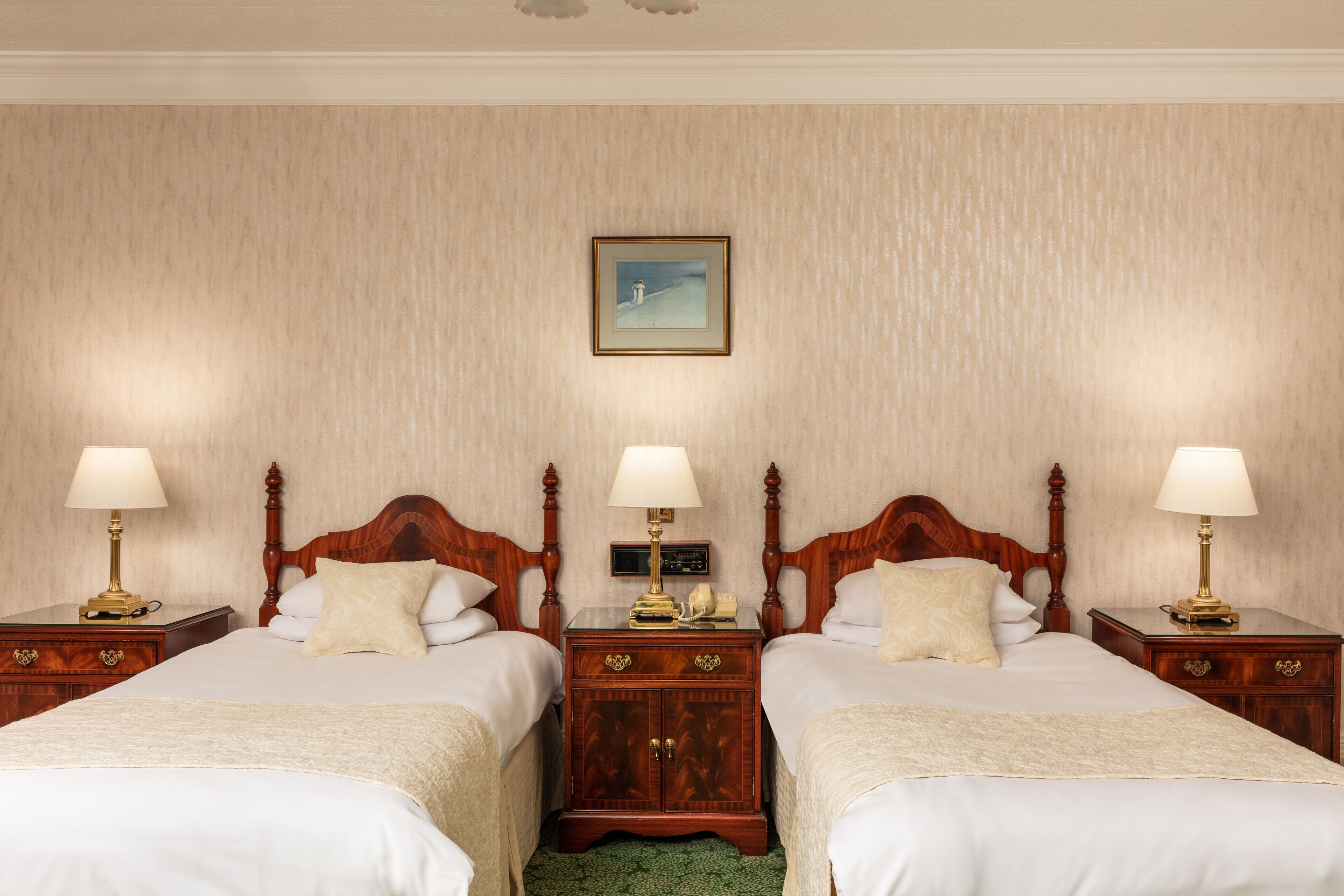 Een hotelkamer met twee bedden, houten hoofdborden, nachtkastjes met lampen, een ingelijst schilderij aan de muur en een groen tapijt.