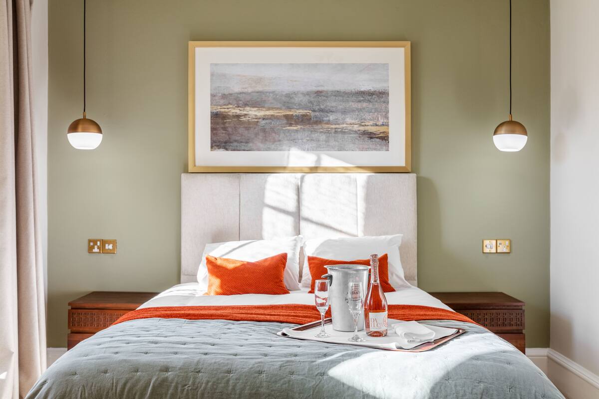 A neatly made bed with a white and orange color scheme, a framed artwork above it, and a bedside table with a bottle and glasses.