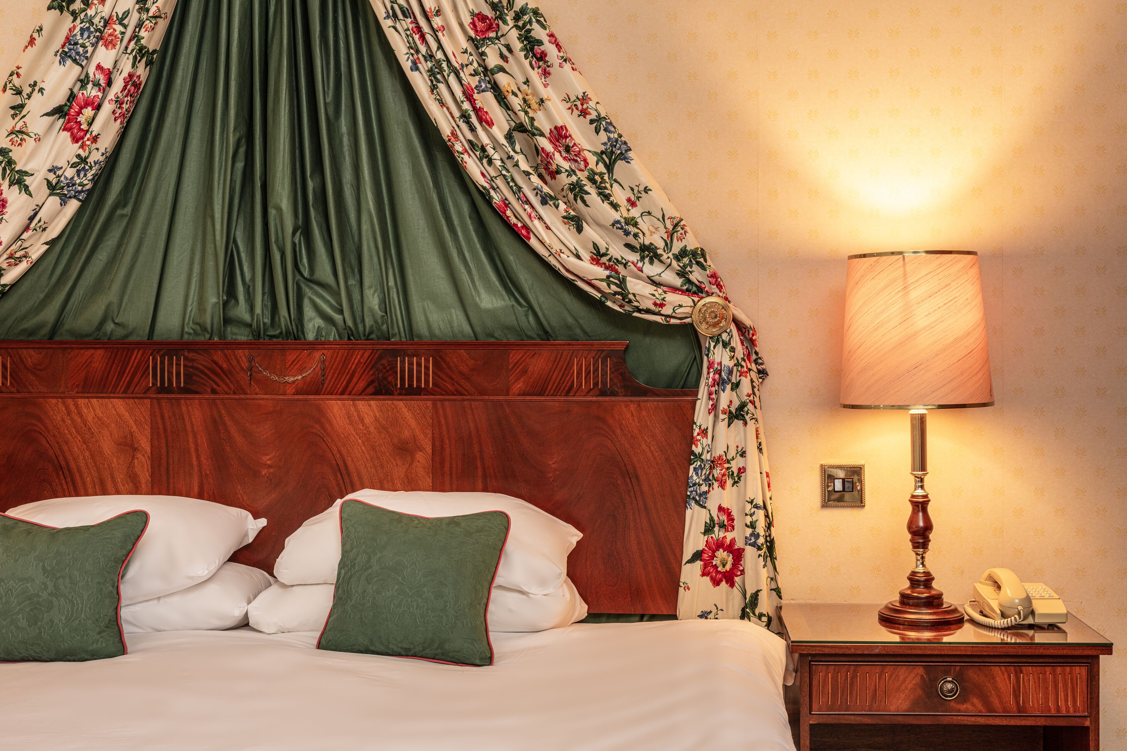 A neatly made bed with green pillows, a wooden headboard, a bedside table with a lamp, and a floral curtain.
