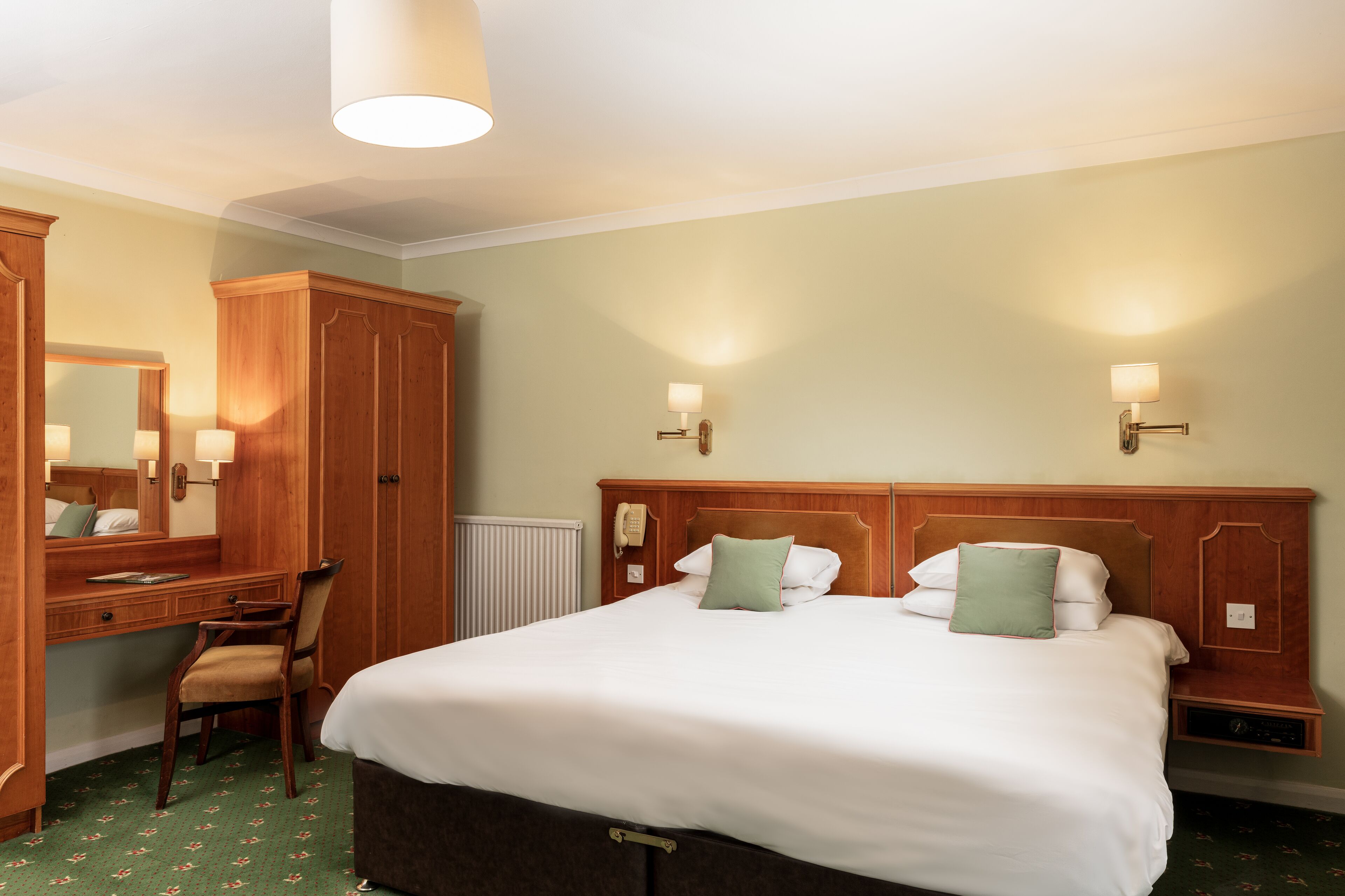A hotel room with a large bed, a wooden wardrobe, a desk with a chair, and two wall-mounted lamps.