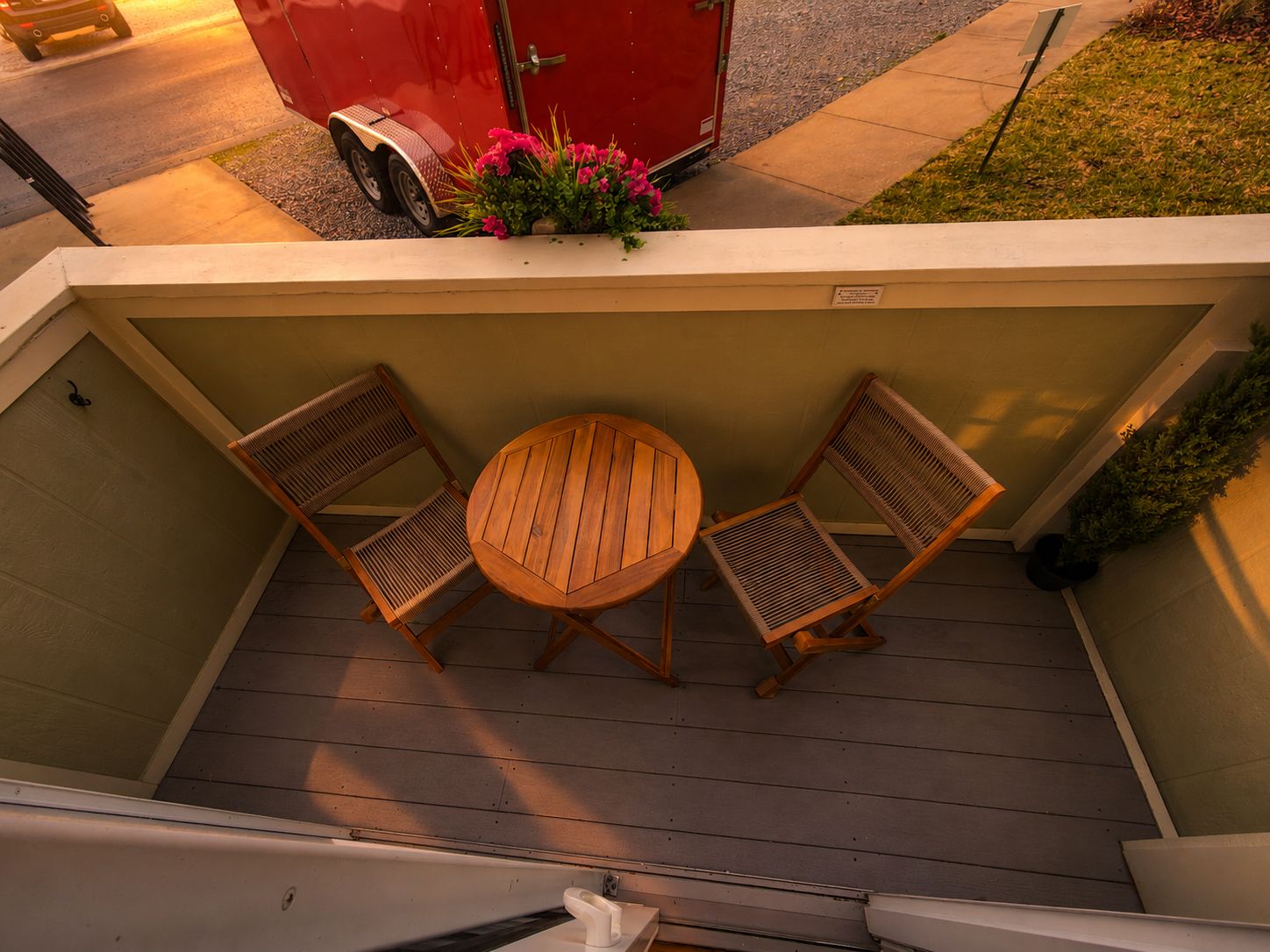 A covered outdoor seating area with a table and two chairs, a red vehicle parked outside, and a flower arrangement on the wall.