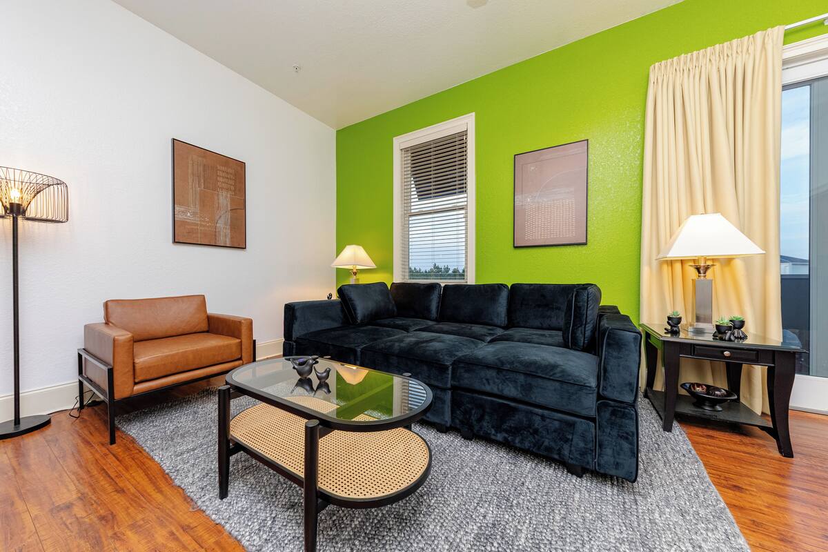 A living room with a black sofa, a brown armchair, and a glass coffee table. There is a lamp on a side table and a window with blinds.