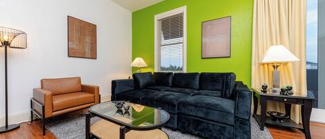 A living room with a black sofa, a brown armchair, and a glass coffee table. There is a lamp on a side table and a window with blinds.