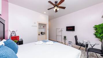Une chambre d’hôtel avec un lit, un ventilateur de plafond, une télévision, un bureau avec des chaises et une petite plante.